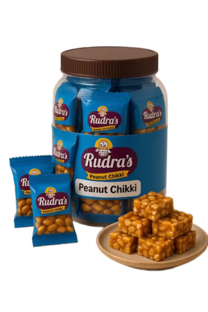 Traditional wooden stove–style presentation of peanut chikki with jaggery-coated groundnut pieces, surrounded by RudrasFoods chikki packets and a small bowl of freshly made peanut chikki samples